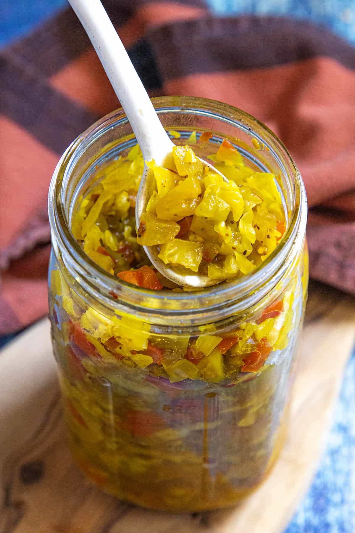 A spoon scoops up chunky, yellow-green Chow Chow Relish from a glass mason jar. The relish contains chopped vegetables, with red and green pieces. A red and black cloth rests in the background on a wooden surface.