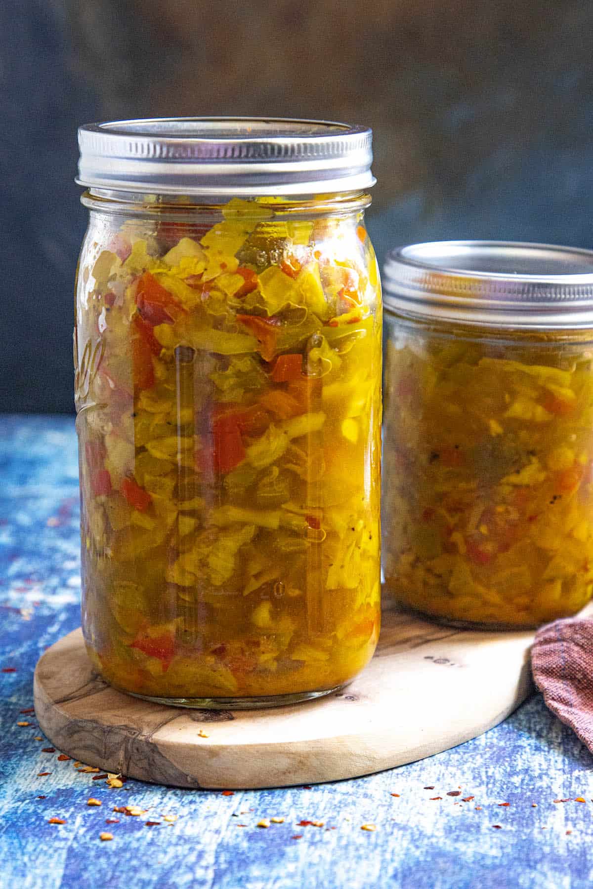 Two glass jars filled with homemade Chow Chow Relish, containing chopped vegetables in brine, sit on a wooden board against a blue background. The jars are sealed with metal lids.