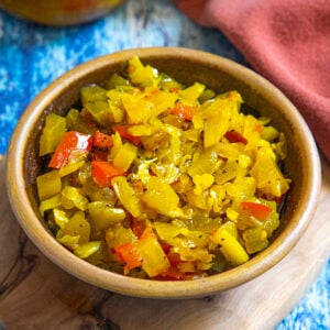 A brown bowl filled with colorful diced vegetables, including yellow, green, and red pieces—likely Chow Chow Relish—sits on a wooden board with a blue and red background.