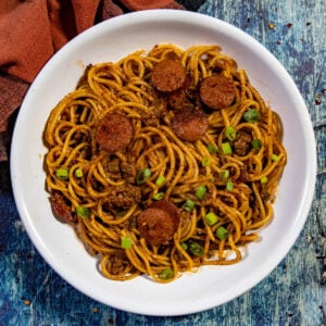 A white bowl filled with Dirty Spaghetti—topped with sliced sausage, ground meat, and garnished with chopped green onions—set on a rustic blue wooden surface with a brown cloth nearby.