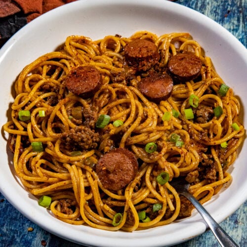 A bowl of Easy-to-Make Dirty Spaghetti mixed with ground meat, sliced sausage, green onions, and red sauce sits with a fork in the bowl. A brown cloth napkin is in the background on a blue textured surface.