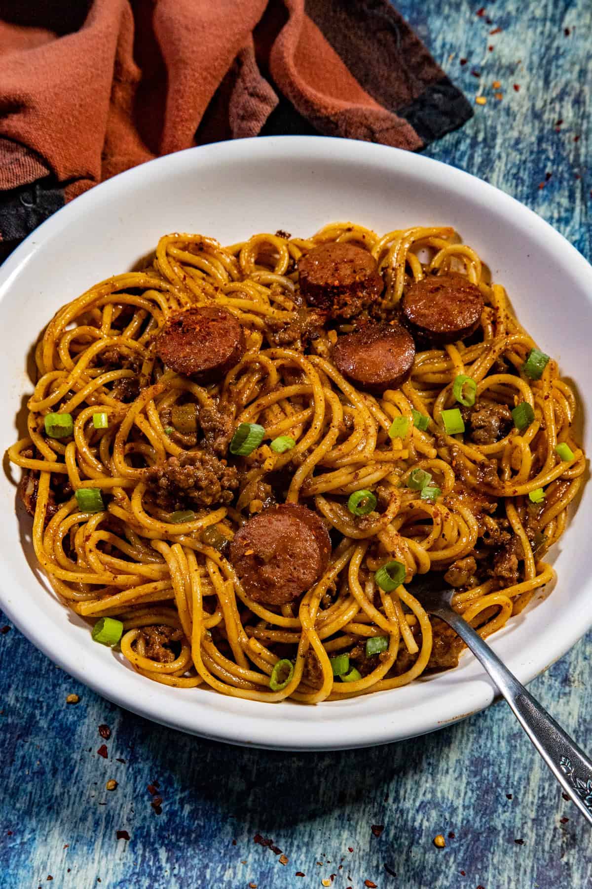 A bowl of Easy-to-Make Dirty Spaghetti mixed with ground meat, sliced sausage, green onions, and red sauce sits with a fork in the bowl. A brown cloth napkin is in the background on a blue textured surface.