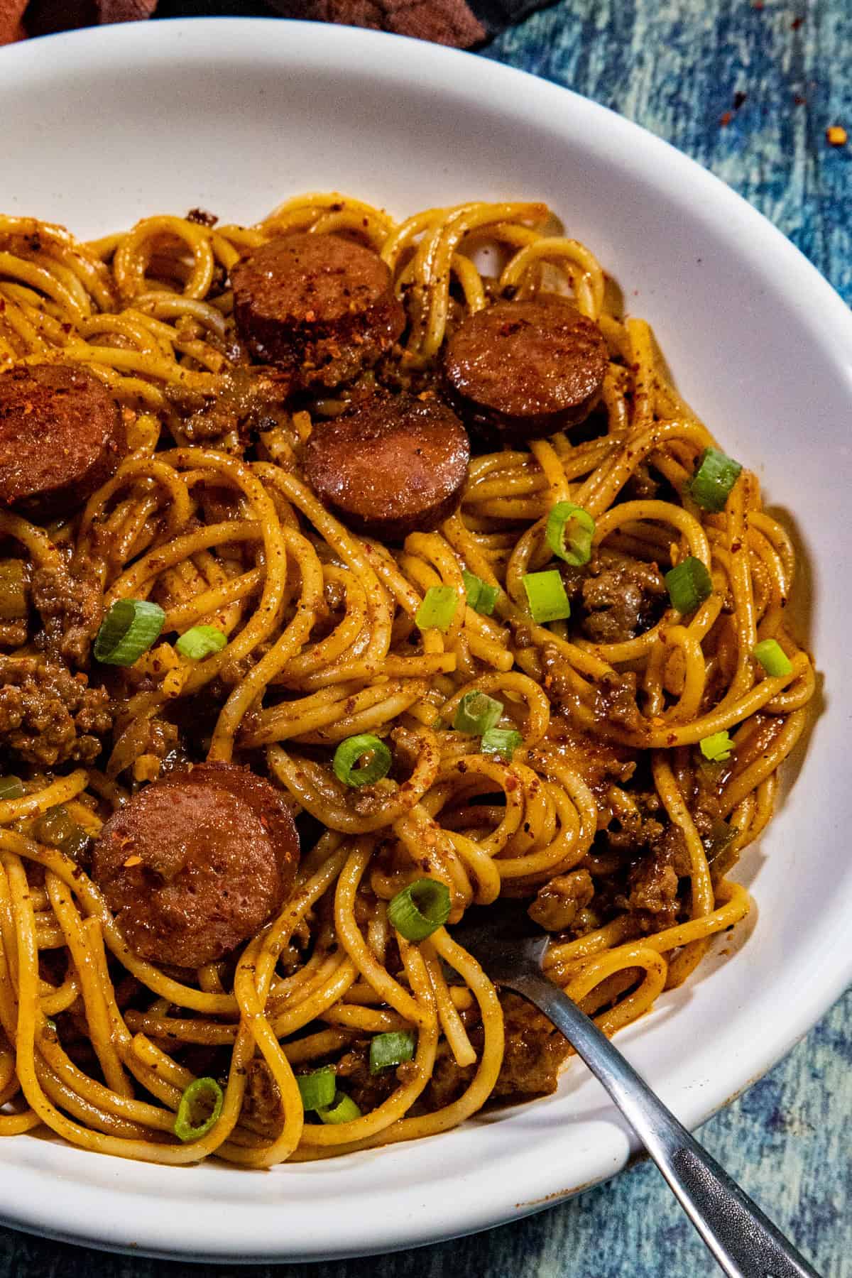 A close-up of Spicy Dirty Spaghetti, topped with sliced sausage, ground meat, and chopped green onions, with a fork resting in the bowl. The dish is served on a white plate against a blue background.