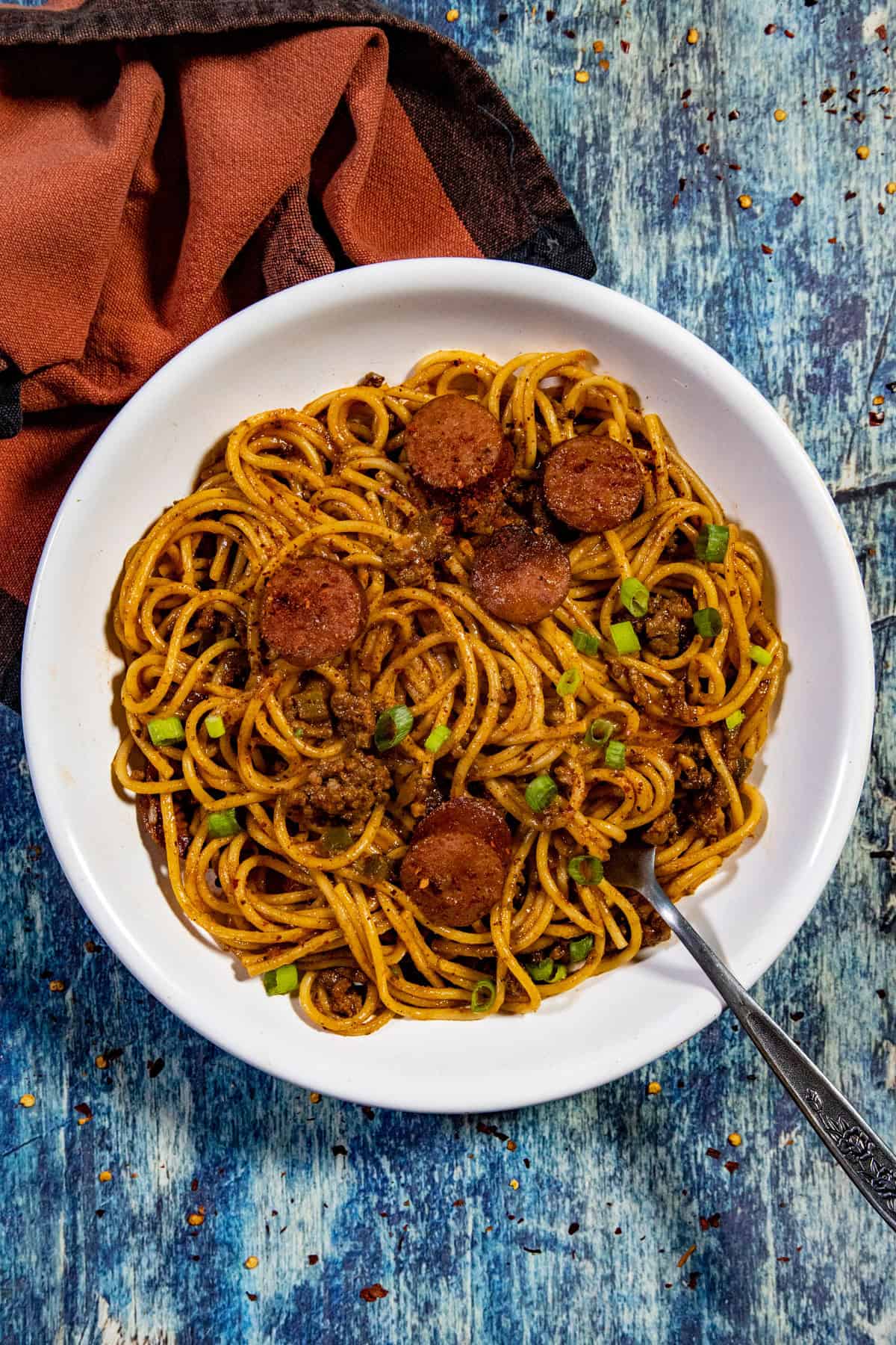 A white bowl filled with Easy Dirty Spaghetti—spaghetti, sliced sausage, ground meat, and chopped green onions—sits on a blue textured surface beside a folded brown napkin. A fork rests in the bowl.
