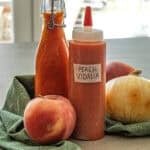 A plastic squeeze bottle labeled Georgia Peach Vidalia Onion Hot Sauce and a glass bottle, both filled with orange sauce, are displayed alongside a peach, a Vidalia onion, and another peach on a green cloth.