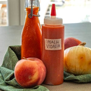 A plastic squeeze bottle labeled Georgia Peach Vidalia Onion Hot Sauce and a glass bottle, both filled with orange sauce, are displayed alongside a peach, a Vidalia onion, and another peach on a green cloth.
