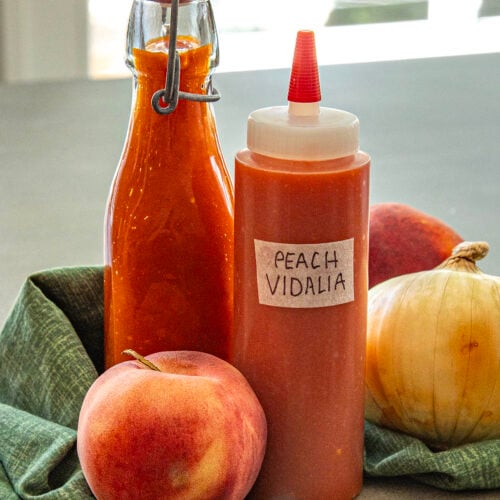 Two bottles of Georgia Peach Vidalia Onion Hot Sauce sit among fresh peaches and a Vidalia onion on a green cloth; one bottle is clear glass with a latch, the other is a squeeze bottle with a handwritten label.