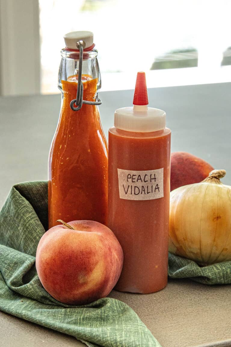 Two bottles of Georgia Peach Vidalia Onion Hot Sauce sit among fresh peaches and a Vidalia onion on a green cloth; one bottle is clear glass with a latch, the other is a squeeze bottle with a handwritten label.