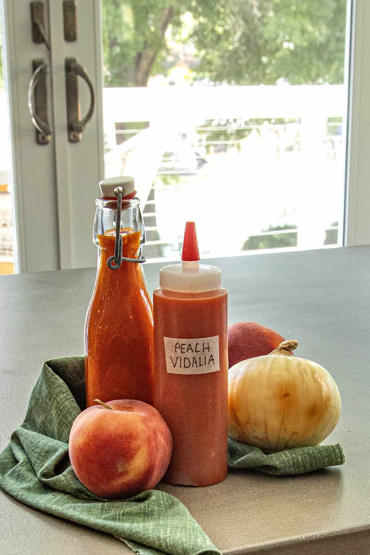 A bottle and a squeeze bottle of Georgia Peach Vidalia Onion Hot Sauce sit on a kitchen counter, surrounded by a fresh peach, Vidalia onion, and green cloth, with sunlight streaming through a bright window in the background.