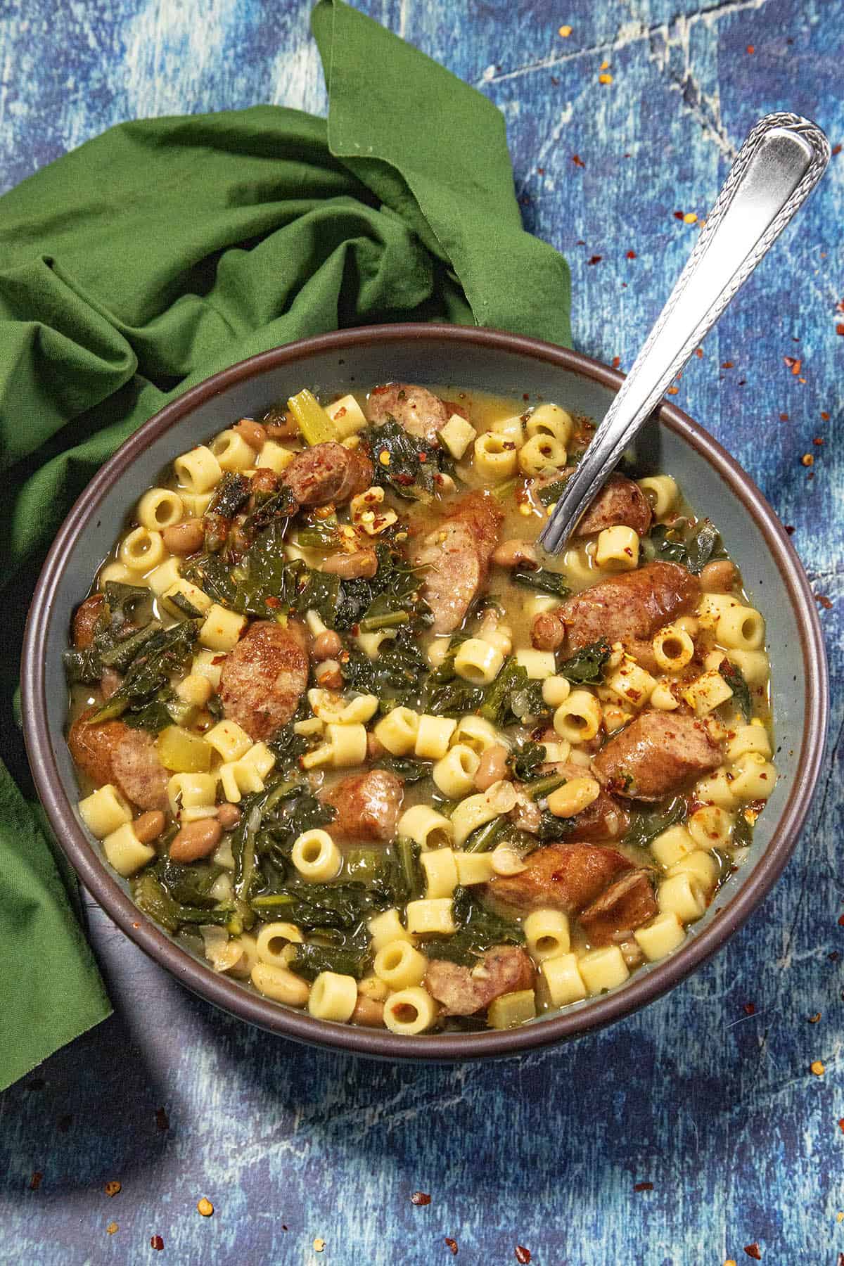 A bowl of pasta soup, reminiscent of Swamp Soup (Turnip Green Soup), with sausage, beans, greens, and ditalini pasta, garnished with red pepper flakes, sits on a blue textured surface next to a green cloth napkin and a spoon in the bowl.