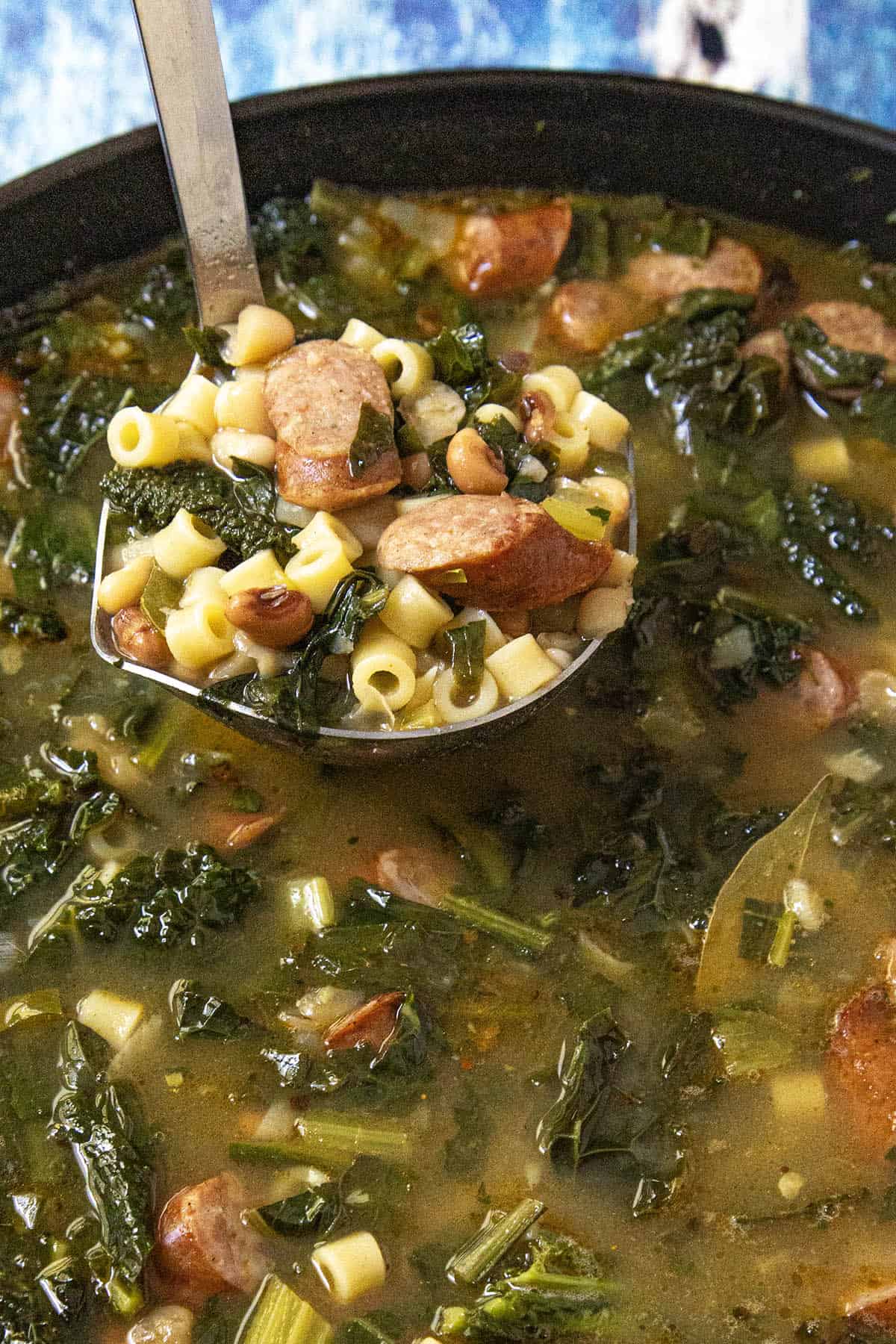 A ladle lifts Swamp Soup with sliced sausage, small pasta, black-eyed peas, and kale from a pot filled with savory broth and greens.