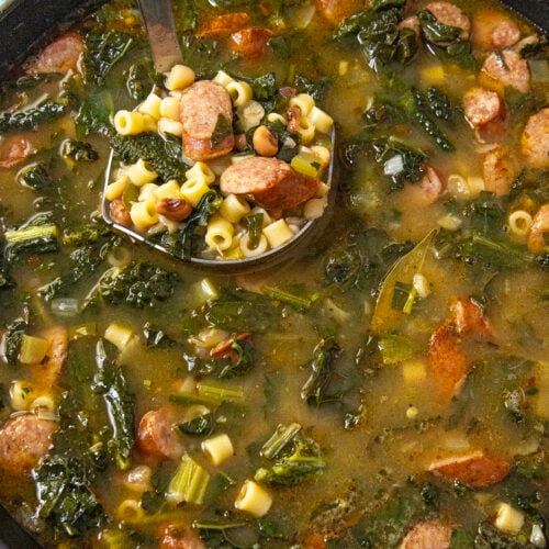 A pot of Swamp Soup (Turnip Green Soup) with sliced sausage, leafy greens, ditalini pasta, and beans. A ladle lifts a serving, showing the ingredients clearly in the savory broth.