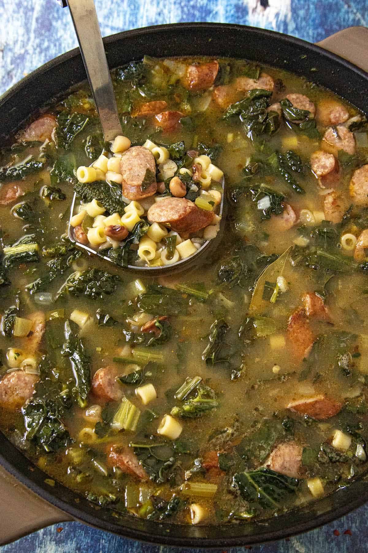 A pot of Swamp Soup (Turnip Green Soup) with sliced sausage, leafy greens, ditalini pasta, and beans. A ladle lifts a serving, showing the ingredients clearly in the savory broth.