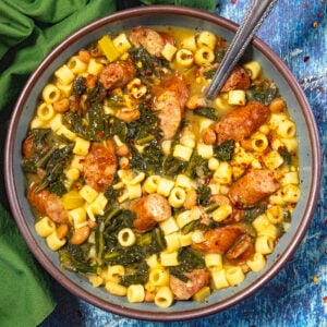 A bowl of Swamp Soup (Turnip Green Soup) with ditalini pasta, sausage slices, beans, leafy greens, and broth, with a spoon in the bowl and a green napkin beside it on a blue textured surface.