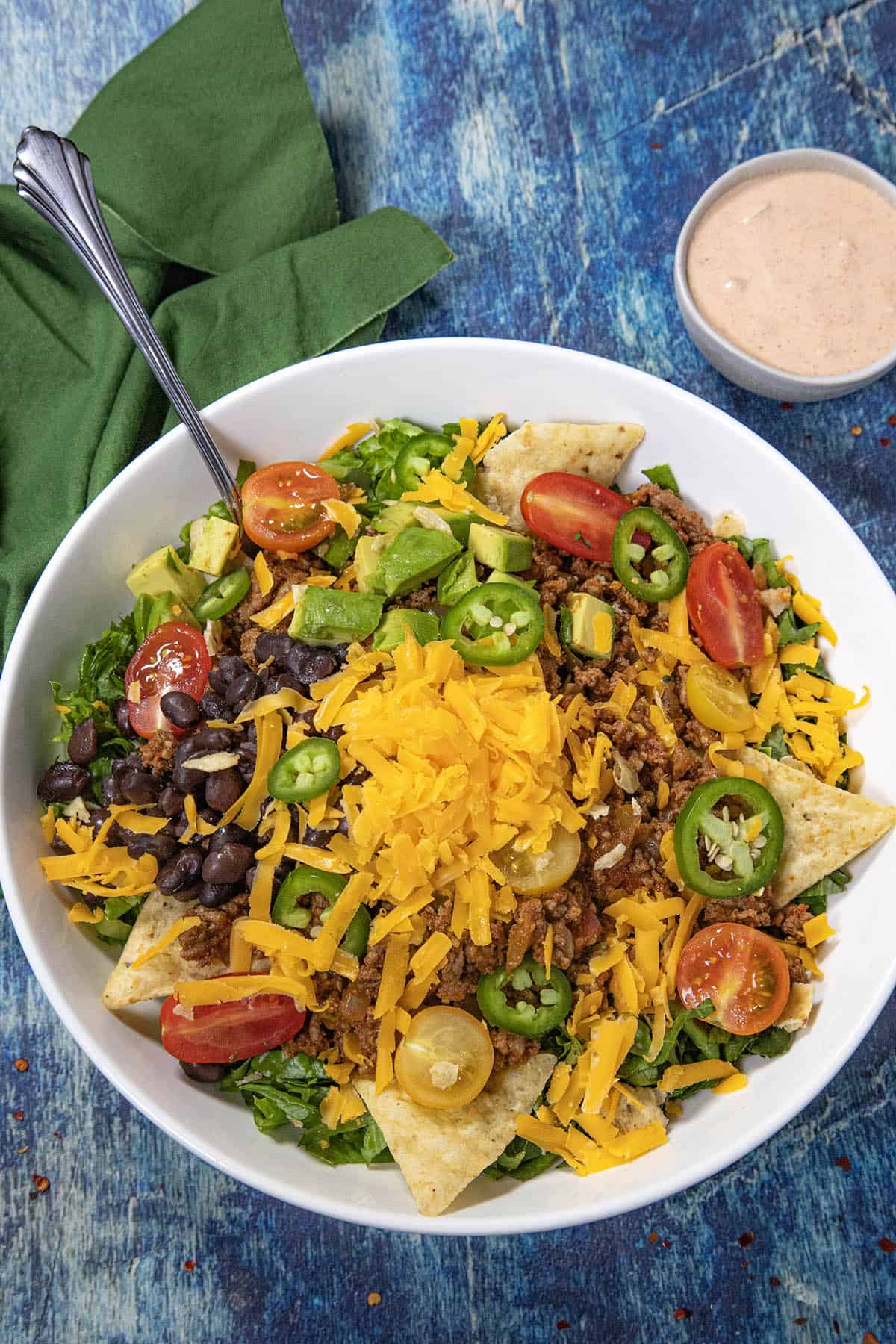 A vibrant Taco Salad in a white bowl topped with cheddar cheese, cherry tomatoes, avocado, black beans, jalapeños, and crisp tortilla chips, served with a fork, green napkin, and creamy dressing on the side.