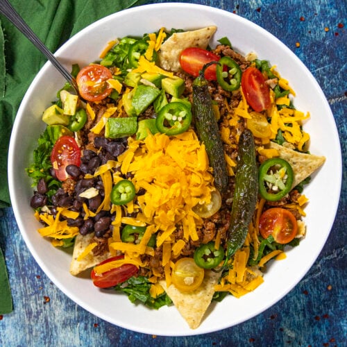 A white bowl filled with vibrant taco salad is topped with shredded cheddar cheese, black beans, tomatoes, jalapeño slices, avocado, tortilla chips, grilled peppers, and crisp lettuce. The colorful taco salad sits on a blue textured surface alongside a green napkin.