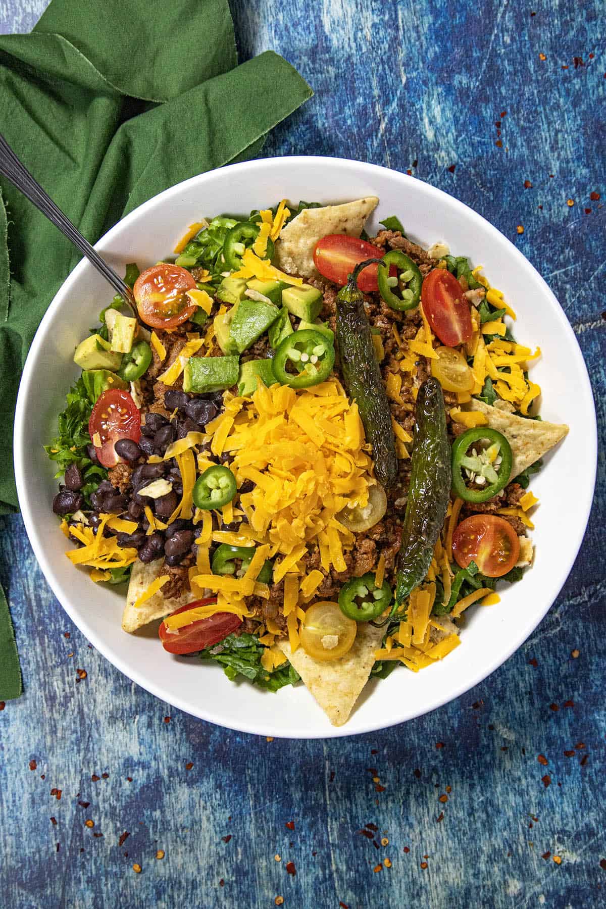 A white bowl filled with vibrant taco salad is topped with shredded cheddar cheese, black beans, tomatoes, jalapeño slices, avocado, tortilla chips, grilled peppers, and crisp lettuce. The colorful taco salad sits on a blue textured surface alongside a green napkin.