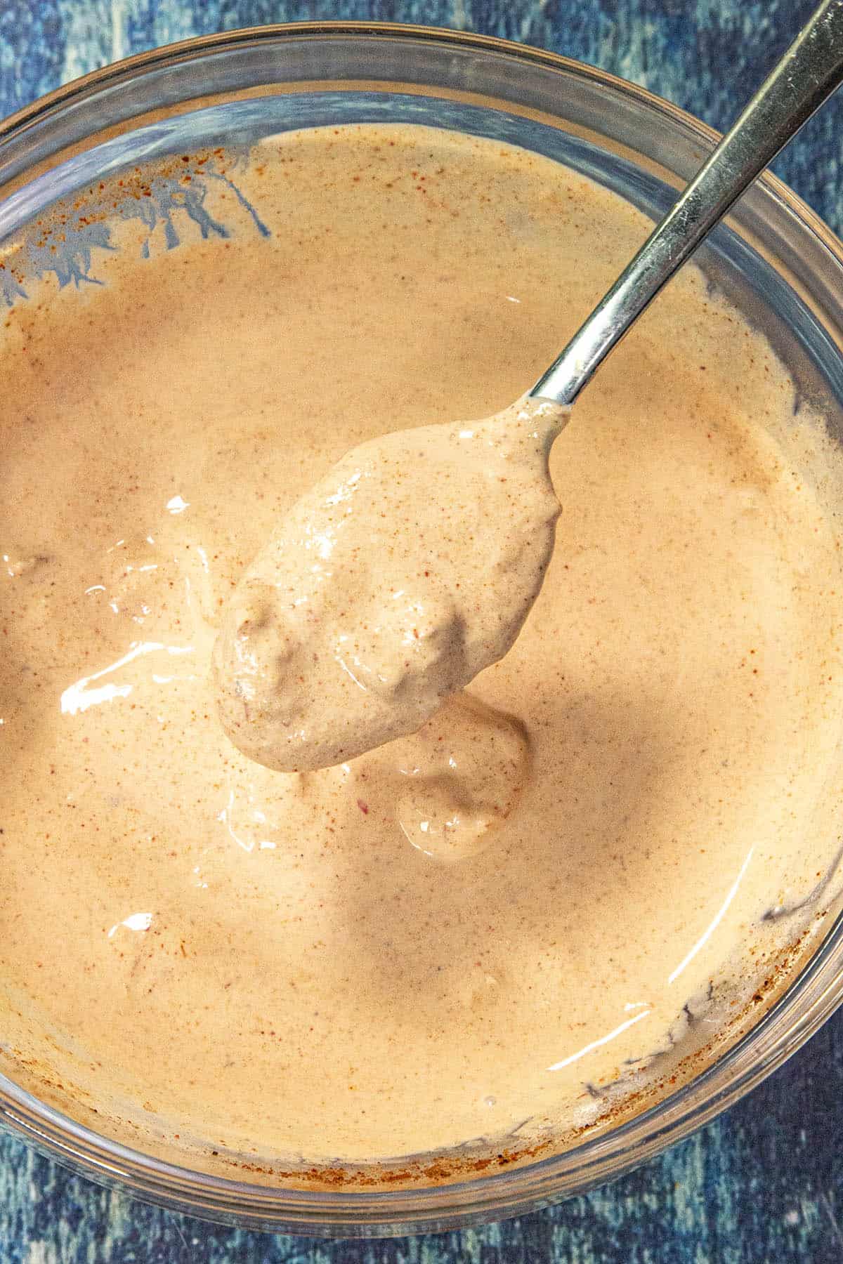 A spoon holds up a thick, creamy taco salad dressing speckled with spices above a clear glass bowl filled with the same taco salad sauce, sitting on a blue textured surface.
