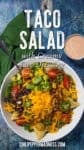 A vibrant Taco Salad bursts with cheddar cheese, ground beef, avocado, tomatoes, black beans, jalapenos, and crunchy tortilla chips, all tossed in a creamy dressing. Served in a bowl with a fork, set alongside a green napkin and a cup of dressing on the side.