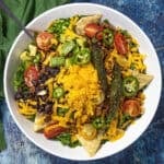 A vibrant Taco Salad in a white bowl with crisp lettuce, savory ground meat, shredded cheddar, black beans, cherry tomatoes, creamy avocado, jalapeños, crunchy tortilla chips, and grilled jalapeños sits on a blue surface, accompanied by a fork and green napkin.