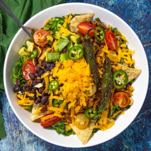 A vibrant Taco Salad in a white bowl with crisp lettuce, savory ground meat, shredded cheddar, black beans, cherry tomatoes, creamy avocado, jalapeños, crunchy tortilla chips, and grilled jalapeños sits on a blue surface, accompanied by a fork and green napkin.