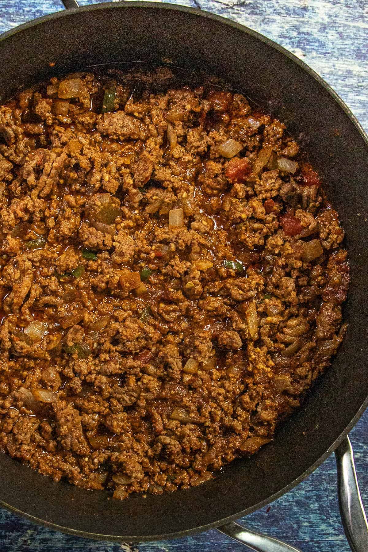 A close-up of a pan filled with cooked ground beef in a rich, chunky tomato sauce with diced onions and peppers. The mixture appears well-seasoned and hearty, making it perfect for tacos, chili, or as a flavorful base for a delicious taco salad.