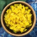 A brown bowl filled with bright yellow tangy Banana Pepper Relish sits on a blue textured surface, with a green cloth partially visible in the corner.