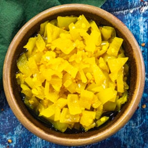 A brown bowl filled with bright yellow tangy Banana Pepper Relish sits on a blue textured surface, with a green cloth partially visible in the corner.
