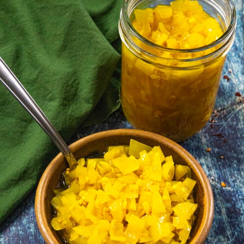 A small bowl and a glass jar filled with bright yellow homemade Banana Pepper Relish sit on a blue surface next to a green cloth. A spoon is in the bowl.