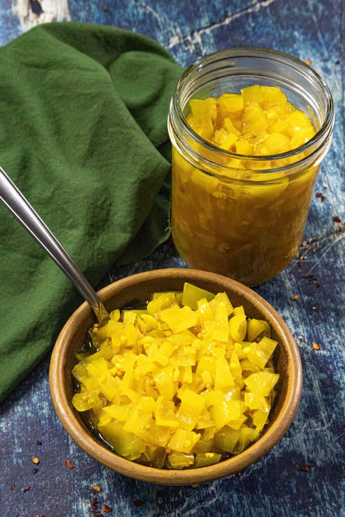 A small bowl and a glass jar filled with bright yellow homemade Banana Pepper Relish sit on a blue surface next to a green cloth. A spoon is in the bowl.
