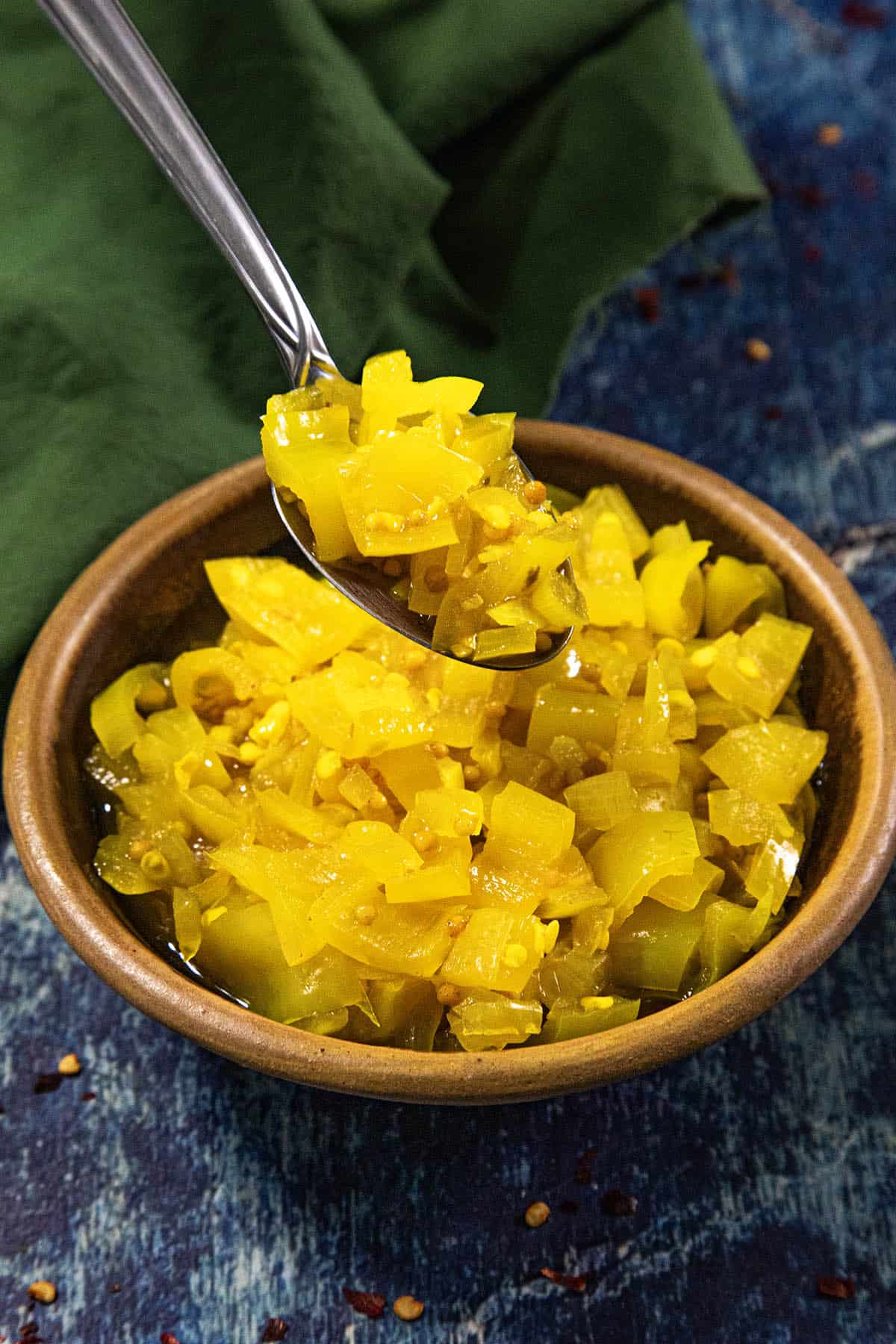 A spoon lifts vibrant yellow Banana Pepper Relish from a brown bowl, which sits on a rustic blue surface with a green cloth nearby.