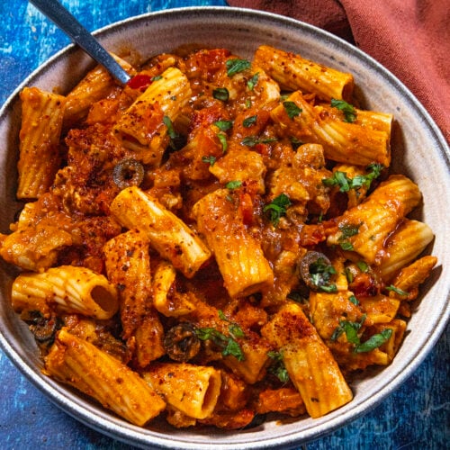 A bowl of Chicken Riggies—rigatoni pasta in a rich tomato sauce with tender chicken, olives, herbs, and spices—sits on a blue surface, with a fork and a brown napkin beside it.