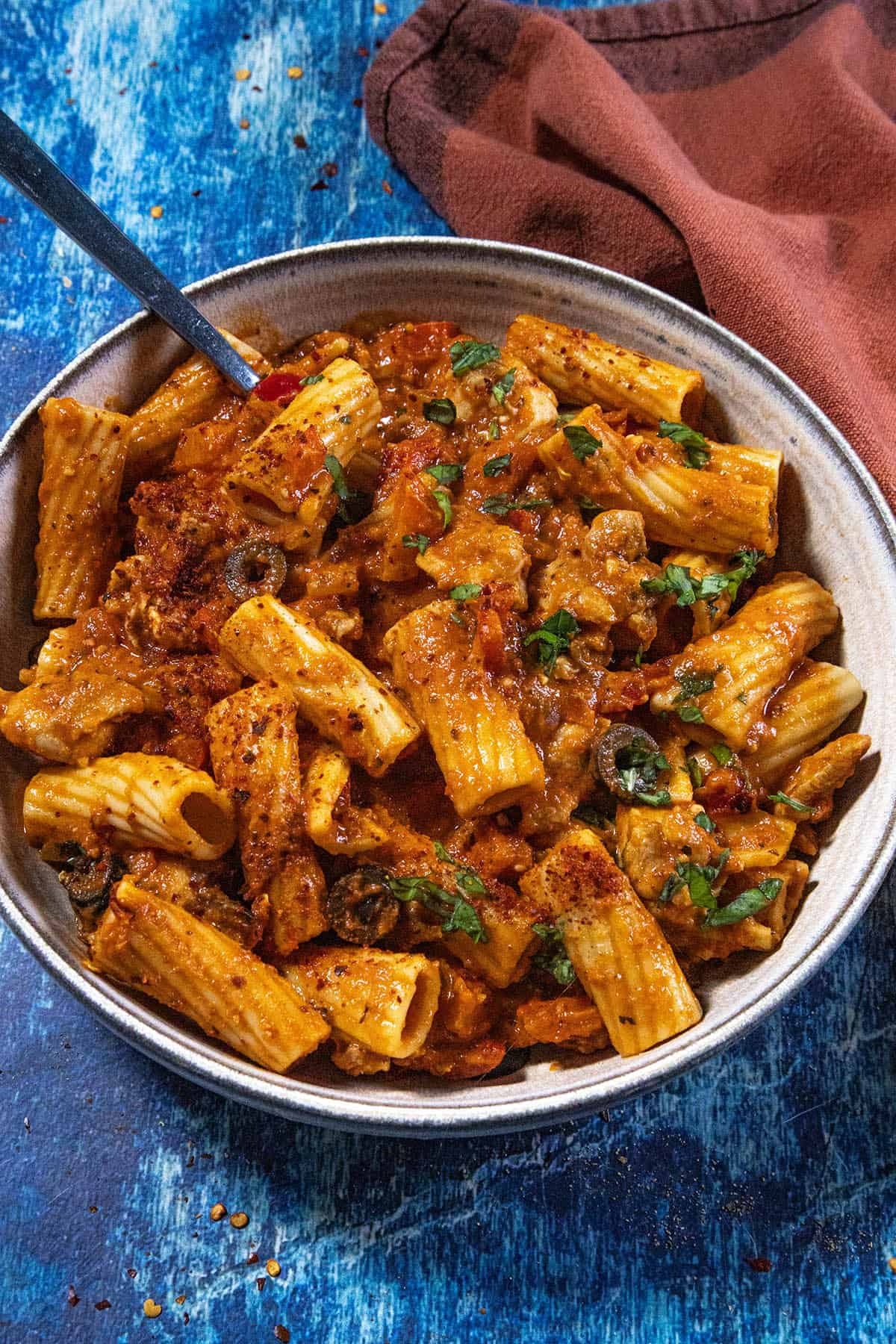 A bowl of Chicken Riggies—rigatoni pasta in a rich tomato sauce with tender chicken, olives, herbs, and spices—sits on a blue surface, with a fork and a brown napkin beside it.
