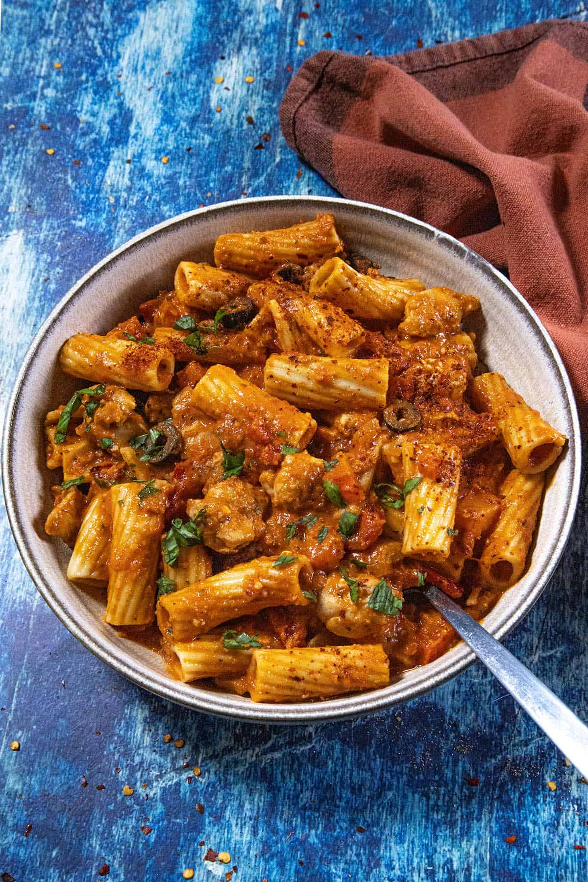 A bowl of Chicken Riggies—rigatoni pasta tossed with tomato sauce, tender chicken, sausage, and fresh herbs—sits on a blue surface next to a brown cloth napkin, with a fork resting in the bowl.