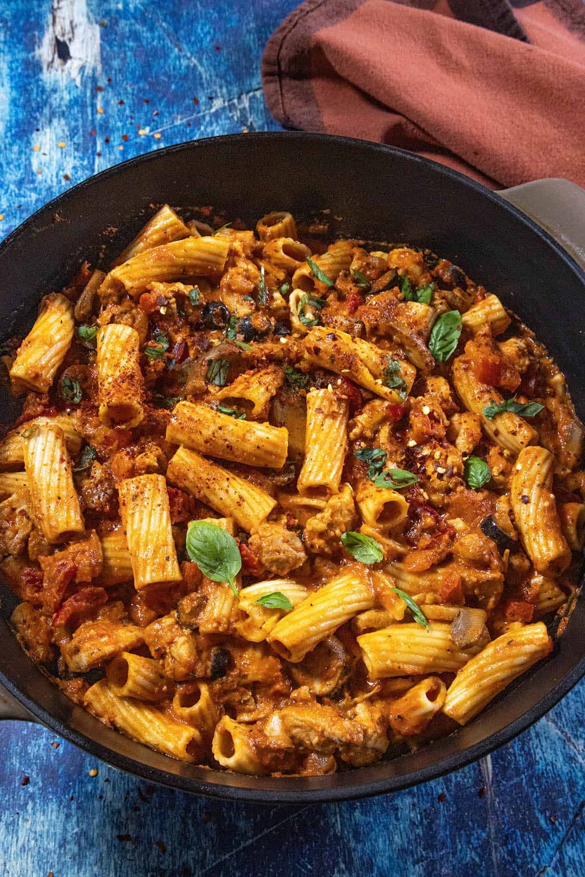 Rigatoni pasta in a skillet with a chunky tomato-based sauce, pieces of cherry peppers, herbs, and garnished with fresh basil leaves— classic Chicken Riggies.