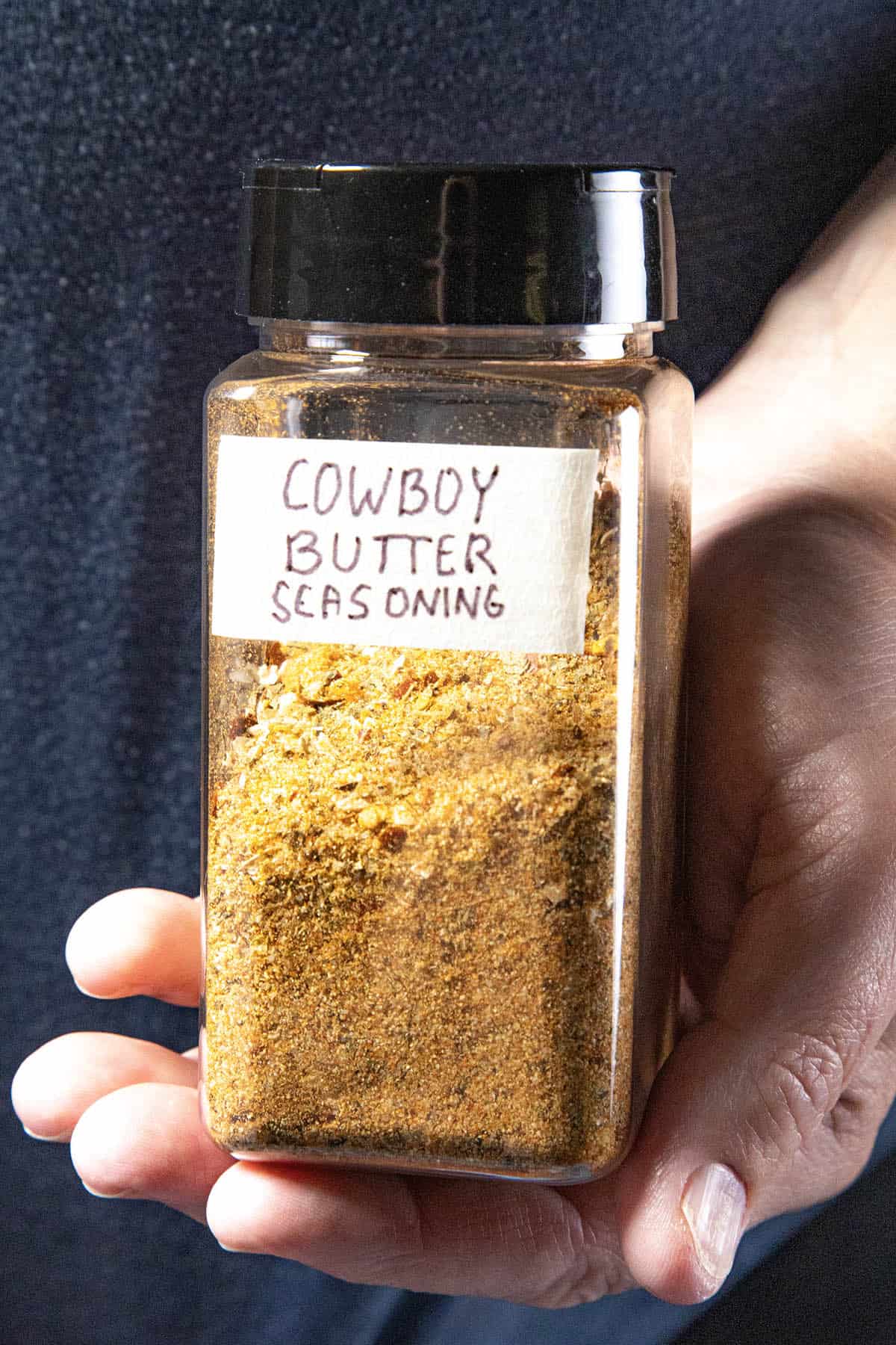 Mike from Chili Pepper Madness holds a clear plastic jar filled with a yellow spice mix labeled Cowboy Butter Seasoning. The jar features a black lid and a handwritten label, highlighting the unique Cowboy Butter Seasoning blend inside.