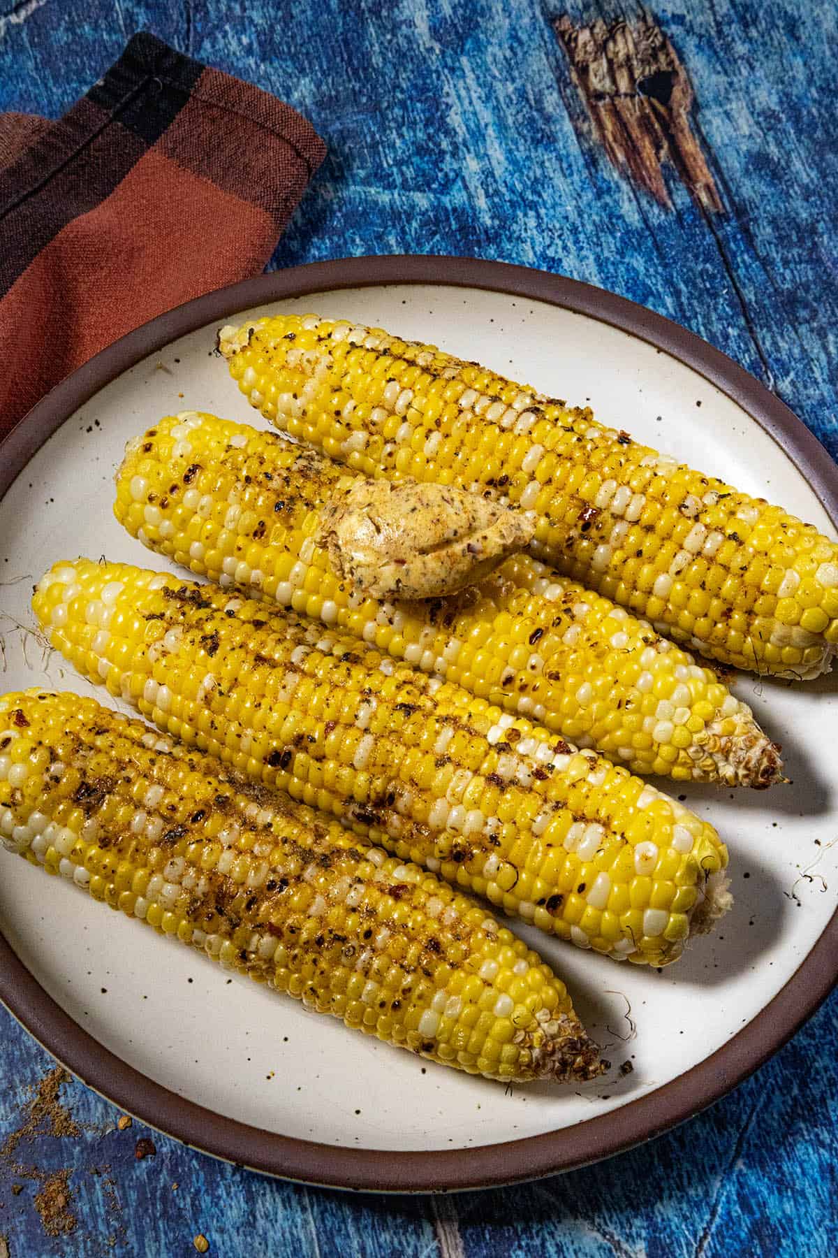 Four grilled corn cobs seasoned with Cowboy Butter Seasoning and topped with a dollop of cowboy butter, served on a white plate with a brown rim, placed on a blue wooden surface with a red and black napkin nearby.