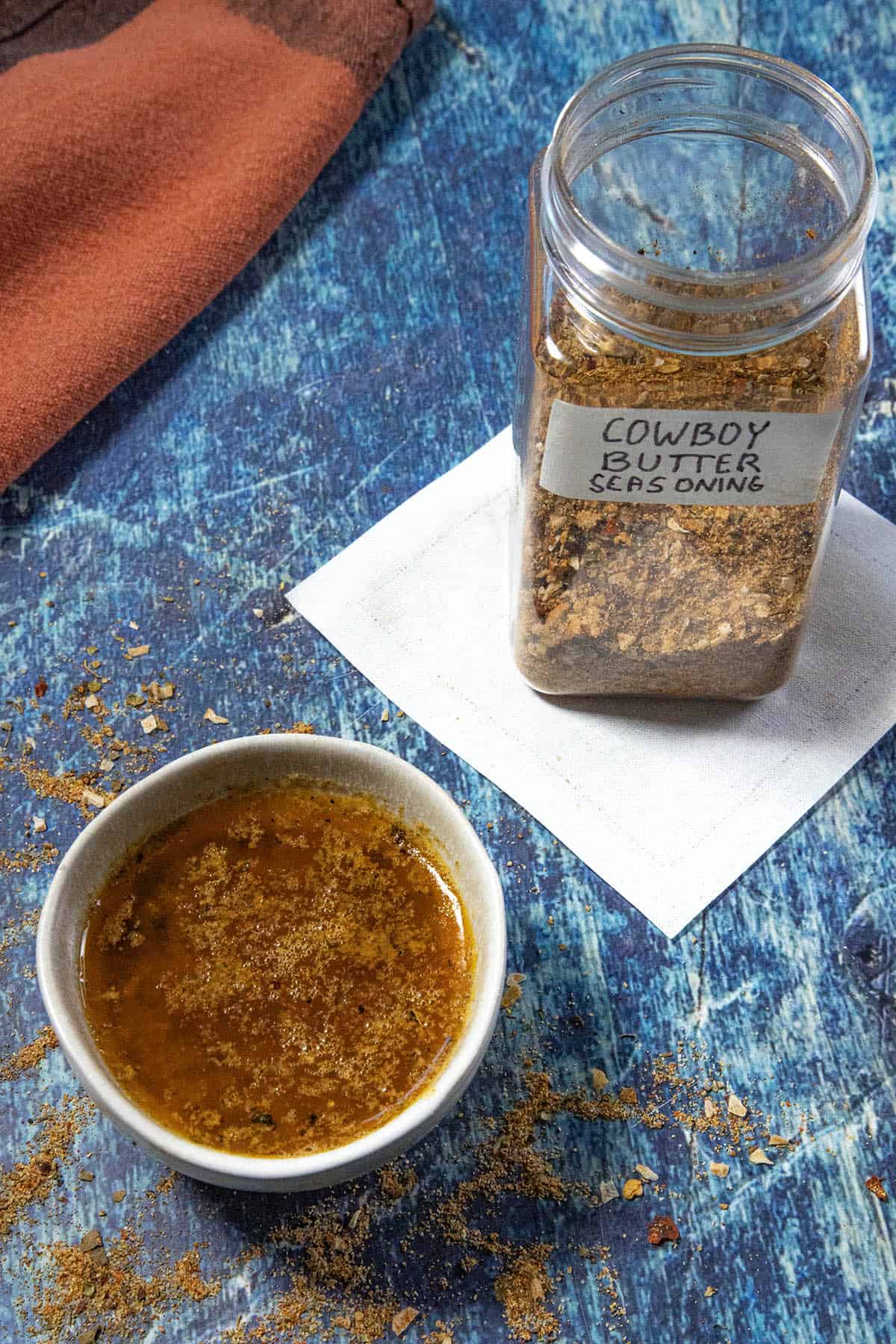 A bowl of Easy Cowboy Butter made from Cowboy Butter Seasoning sits beside a labeled jar of dry cowboy butter spices on a blue surface, with some seasoning scattered around.