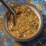 A bowl filled with a coarse, mixed spice blend containing herbs and crushed chili flakes—reminiscent of Cowboy Butter Seasoning—with a spoon resting inside, on a blue textured surface.