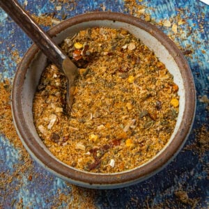 A bowl filled with a coarse, mixed spice blend containing herbs and crushed chili flakes—reminiscent of Cowboy Butter Seasoning—with a spoon resting inside, on a blue textured surface.
