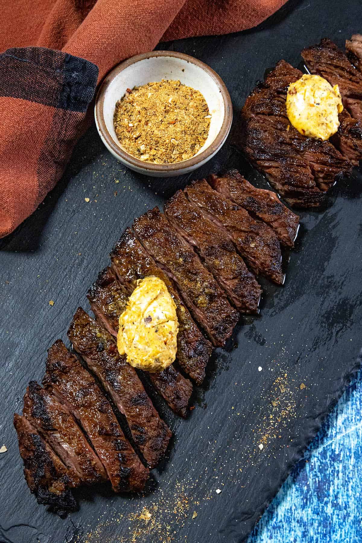 Sliced grilled steak topped with dollops of seasoned Cowboy Butter, served on a black slate with a bowl of dry cowboy butter seasoning. A red cloth and blue fabric are partially visible in the background.