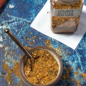 A bowl of Cowboy Butter Seasoning with a spoon sits on a blue surface. Behind it is a labeled jar filled with the same Cowboy Butter Seasoning blend, resting on a white paper.
