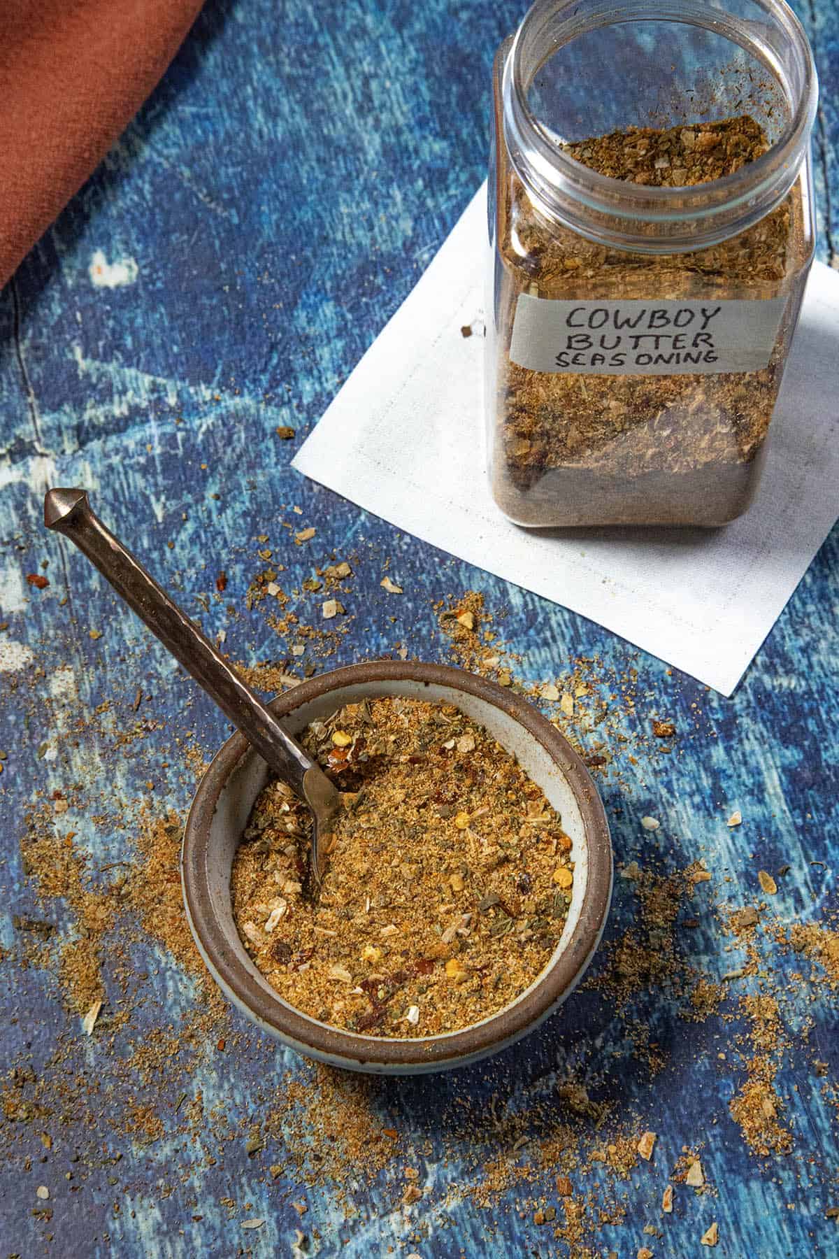 A bowl of Cowboy Butter Seasoning with a spoon sits on a blue surface. Behind it is a labeled jar filled with the same Cowboy Butter Seasoning blend, resting on a white paper.