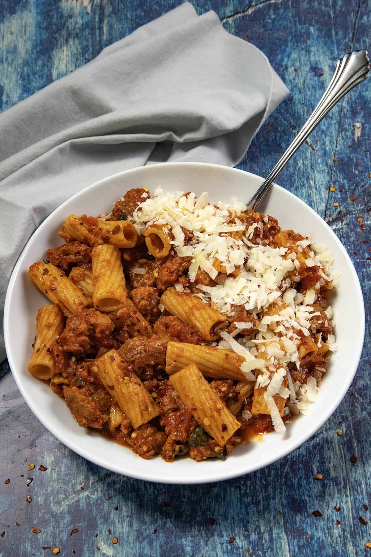 A white bowl of Spicy Italian Sausage Pasta with meat sauce, topped with grated cheese, sits on a blue textured surface with a silver fork and a gray cloth napkin nearby.