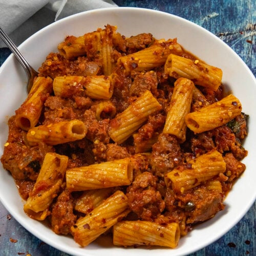 A white bowl filled with spicy Italian sausage pasta and rigatoni in a rich tomato sauce sits on a blue surface, with a fork partially visible beside a gray napkin.