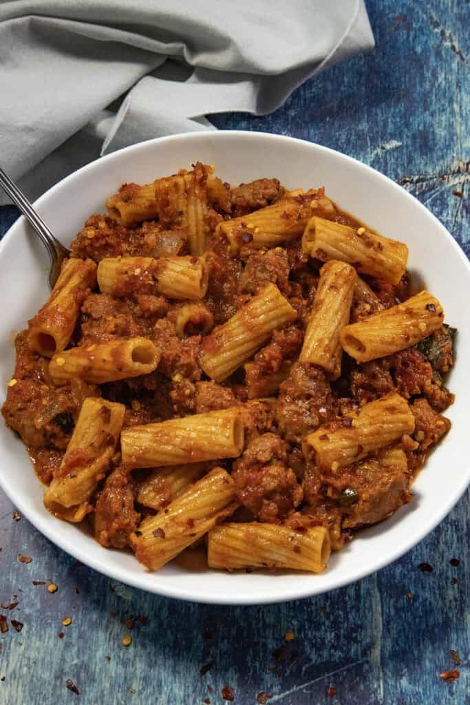A white bowl filled with spicy Italian sausage pasta and rigatoni in a rich tomato sauce sits on a blue surface, with a fork partially visible beside a gray napkin.