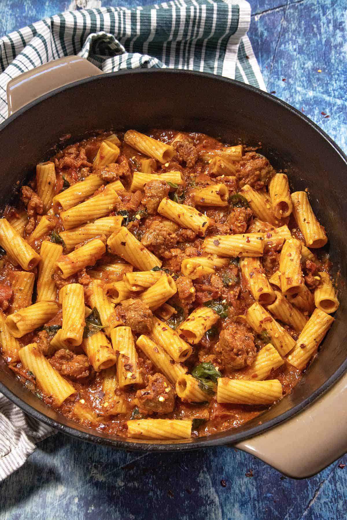 Cooked rigatoni pasta mixed with a rich tomato sauce and chunks of Italian sausage in a large black pot, with a striped kitchen towel underneath, set on a blue textured surface—bringing you the comforting flavors of classic Italian Sausage Pasta.