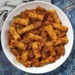 A white bowl filled with Italian Sausage Pasta—rigatoni mixed with flavorful ground meat and tomato sauce, garnished with red pepper flakes—sits on a blue textured surface.