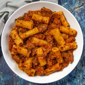 A white bowl filled with Italian Sausage Pasta—rigatoni mixed with flavorful ground meat and tomato sauce, garnished with red pepper flakes—sits on a blue textured surface.