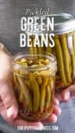 A person holds two glass jars filled with Pickled Green Beans, spices, and brine. The text reads Pickled Green Beans and chilipeppermadness.com.